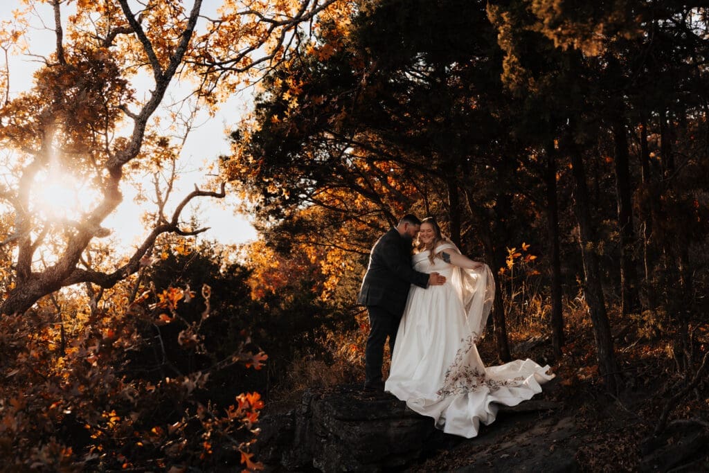 Photo of Rebecca & Justin in their wedding attire surrounded by big beautiful trees that have turned colors for Fall.