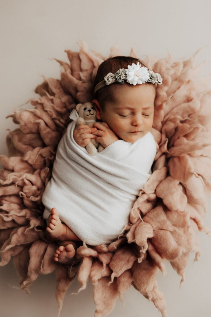 Newborn baby Leonie wrapped in a white wrap, with a floral headpiece on top of a pink fluffy textured fur piece with a cute little teddy bear in her hands.
