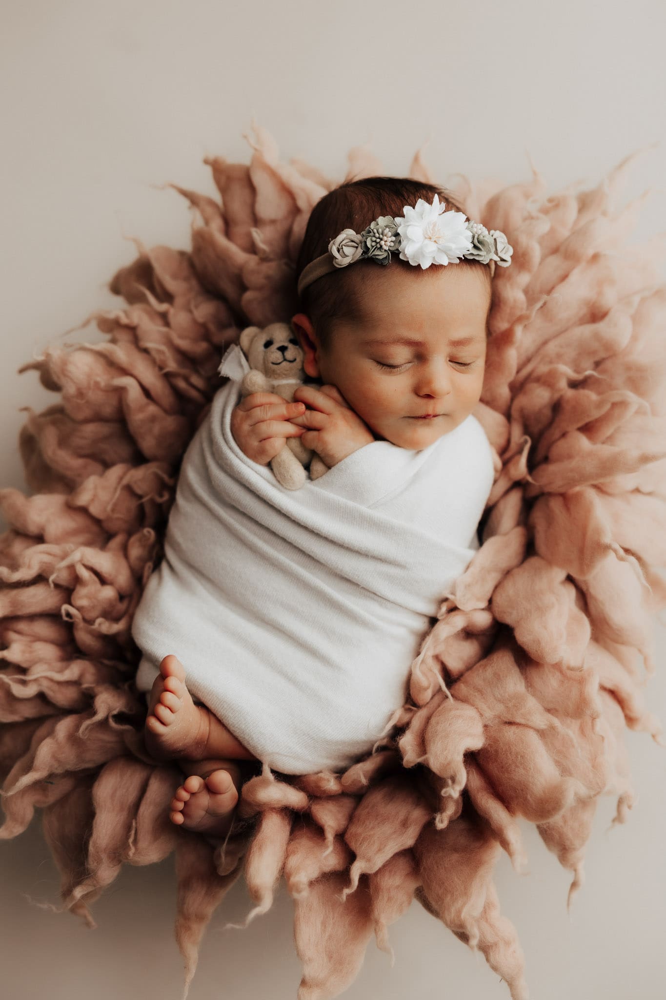 Newborn baby Leonie wrapped in a white wrap, with a floral headpiece on top of a pink fluffy textured fur piece with a cute little teddy bear in her hands.
